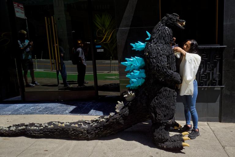 A person helping someone in a godzilla costume drink water. The godzilla costume is loooong