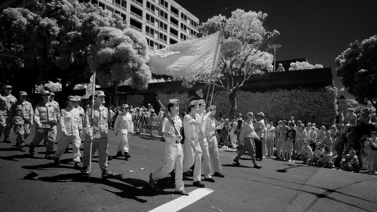 Black and white infrared photograph of marching ROTC folks with protestors looking on from the side. Part of a banner is visible, 