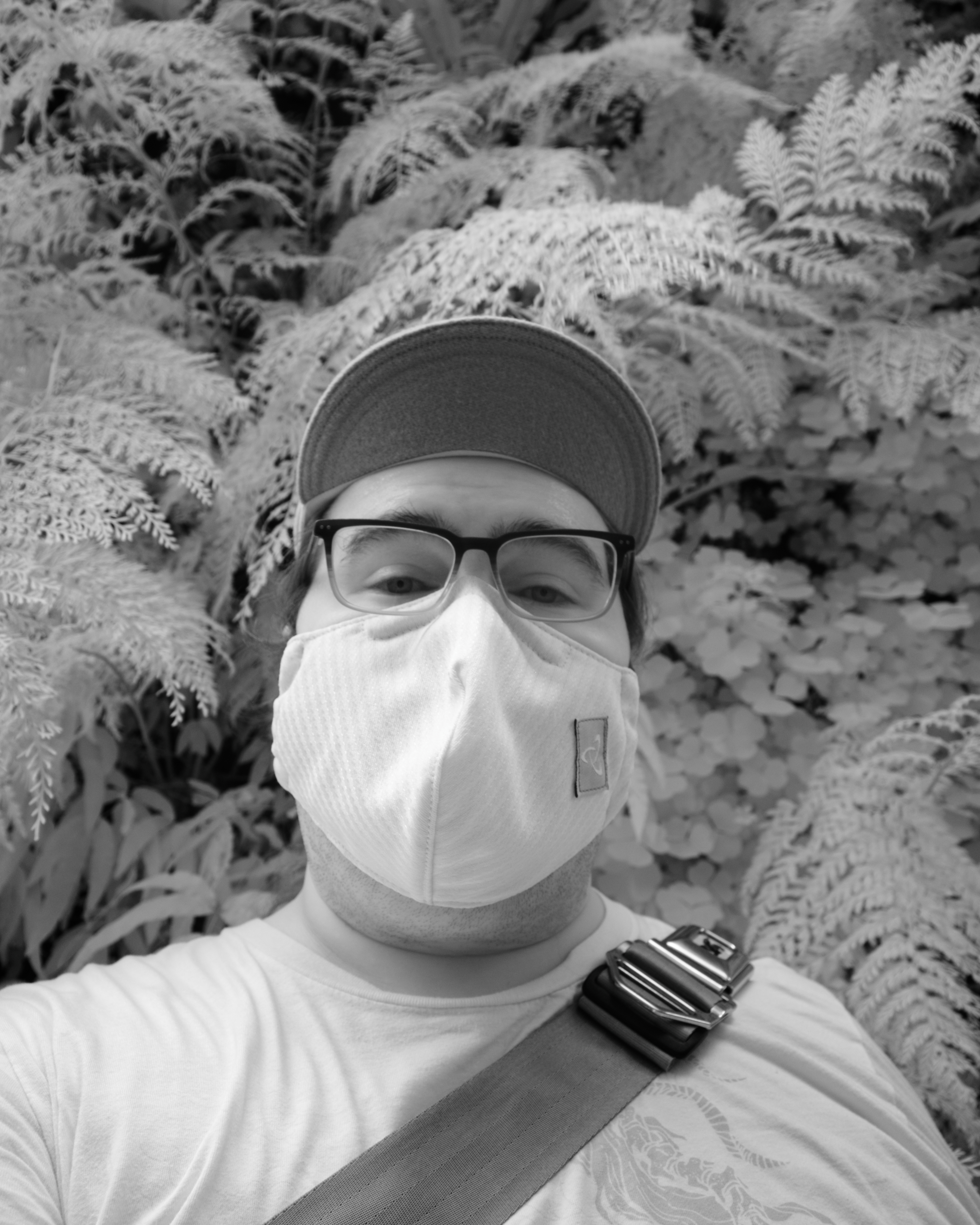 Black and white infrared photograph of a person wearing a mask, a baseball cap, and glasses, standing in front of a green wall.