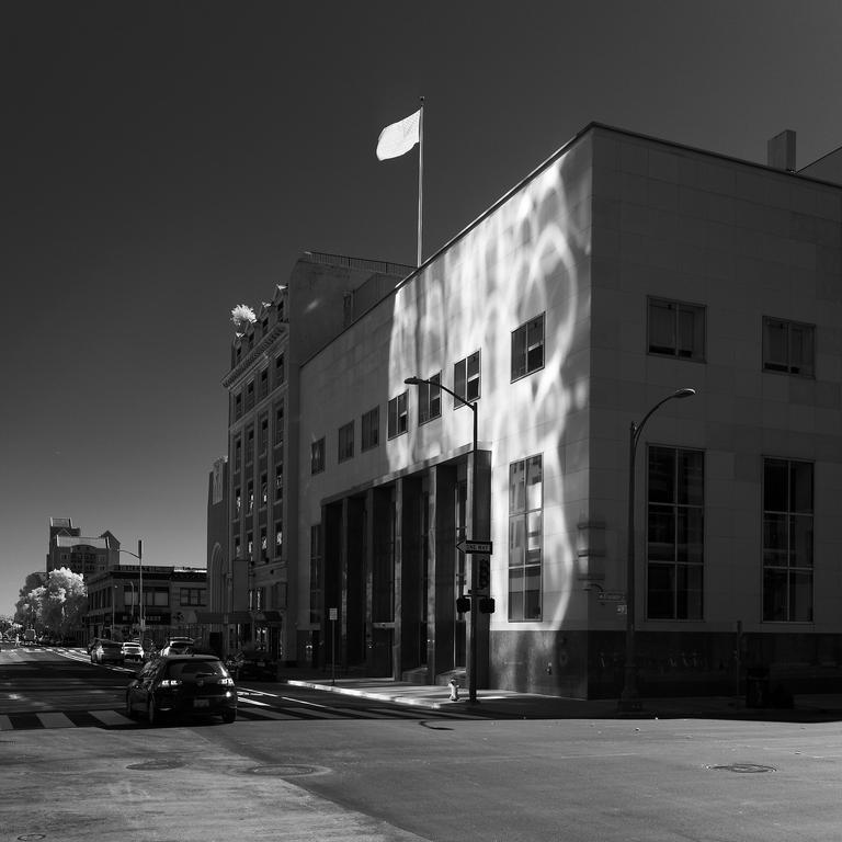 Black and white infrared photo of a 3-4 story cuboid building with a flagpole on top. The side of the building is illuminated with many overlapping circular/oblong pools of light, creating a somewhat spirographic effect. The US flag on the building appears as almost featureless