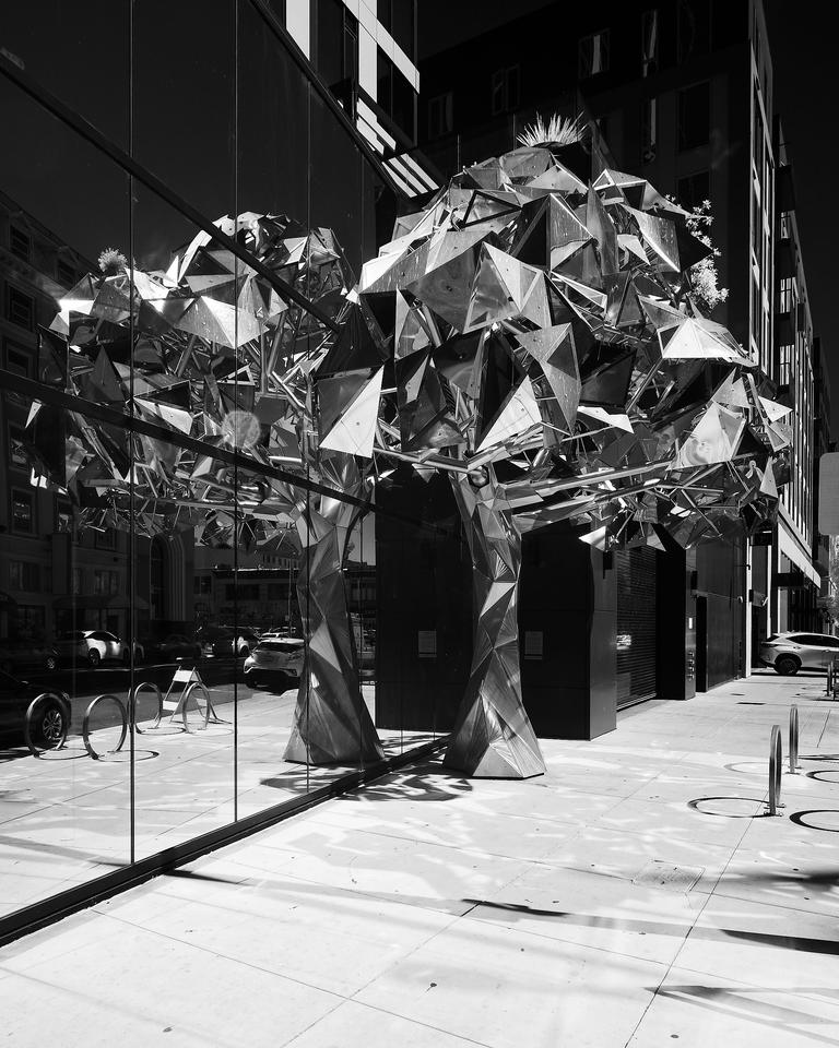 Black and white infrared photo of a metal sculpture depicting a tree. It is composed of many flat metal pieces assembled together and stands in front of a glass office building.