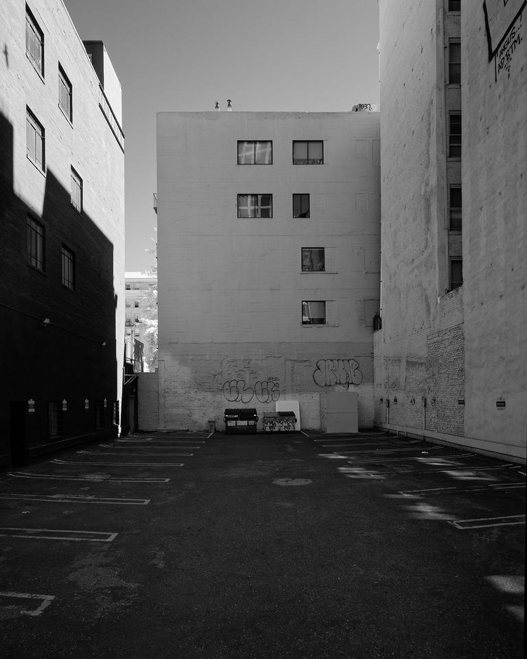 An empty parking lot surrounded by three tall buildings in deep shade. A tree can be seen peeking around the corner of one building. 