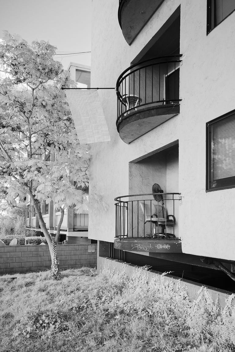 Black and white infrared photograph of a large US flag displayed from the second floor balcony of an apartment. The flag's most visible features are creases. 