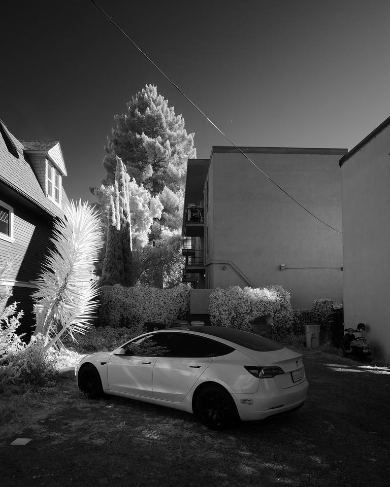 Black and white infrared photo of a small parking lot amongst small apartment buildings, framed by bushes, trees, and a utility wire crossing the sky above a parked car. 